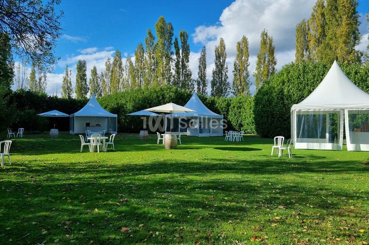 Espace extérieur avec pelouse, tentes blanches, tables et chaises sous un ciel partiellement nuageux.