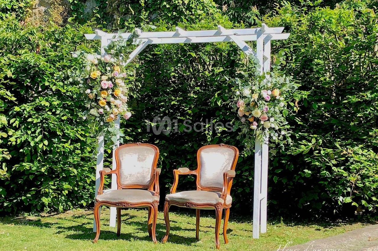 Deux chaises anciennes sous une arche blanche décorée de fleurs, placées dans un jardin verdoyant.