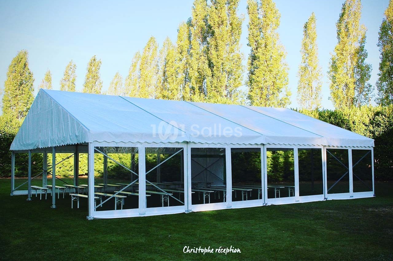 Grande tente blanche installée sur une pelouse, entourée d'arbres, avec des parois transparentes.