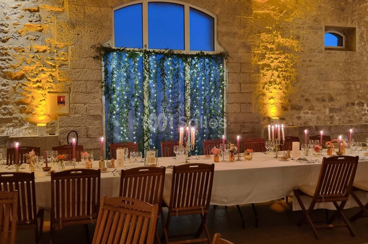 Grande table dressée pour un dîner, décorée de chandeliers et guirlandes lumineuses, dans une salle en pierre éclairée.
