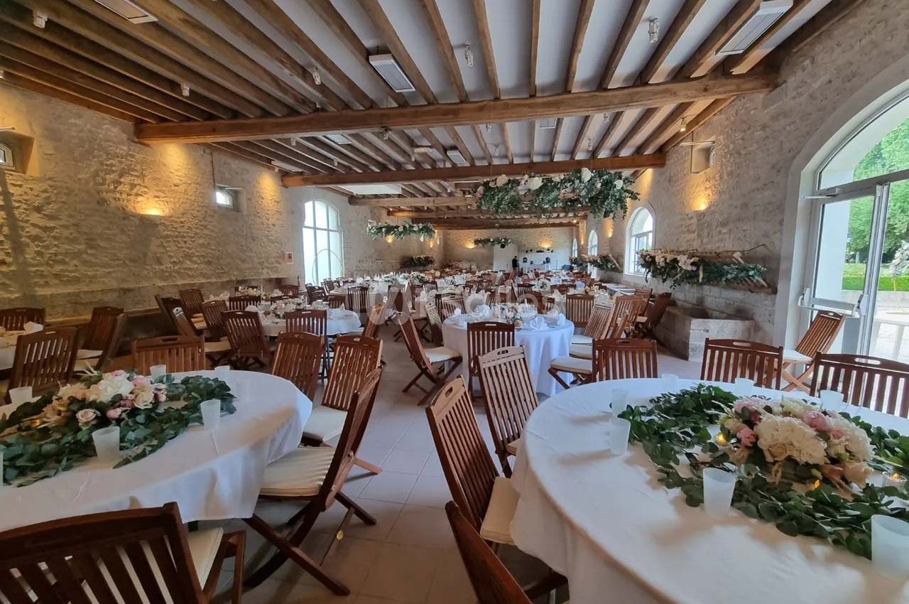 Salle de réception lumineuse avec tables rondes décorées de fleurs, chaises en bois et poutres apparentes au plafond.