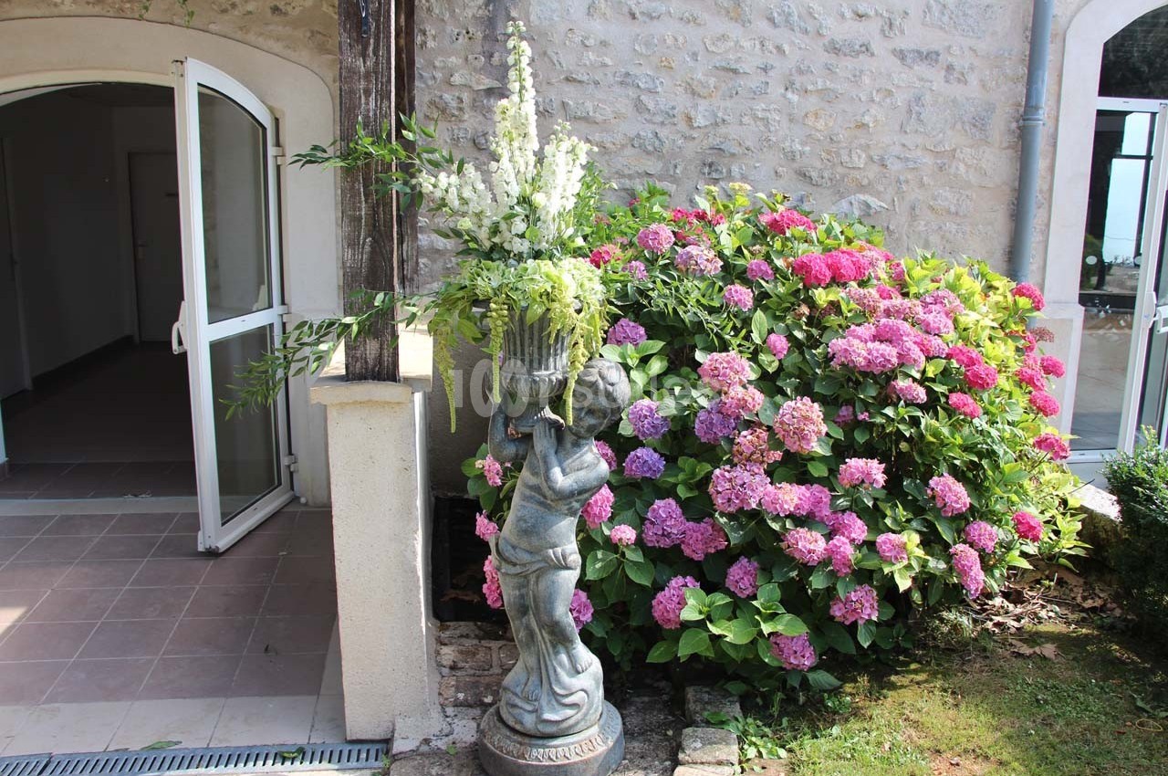Statue décorative portant un vase fleuri devant un mur en pierre, près d'hortensias roses et d'une porte ouverte.