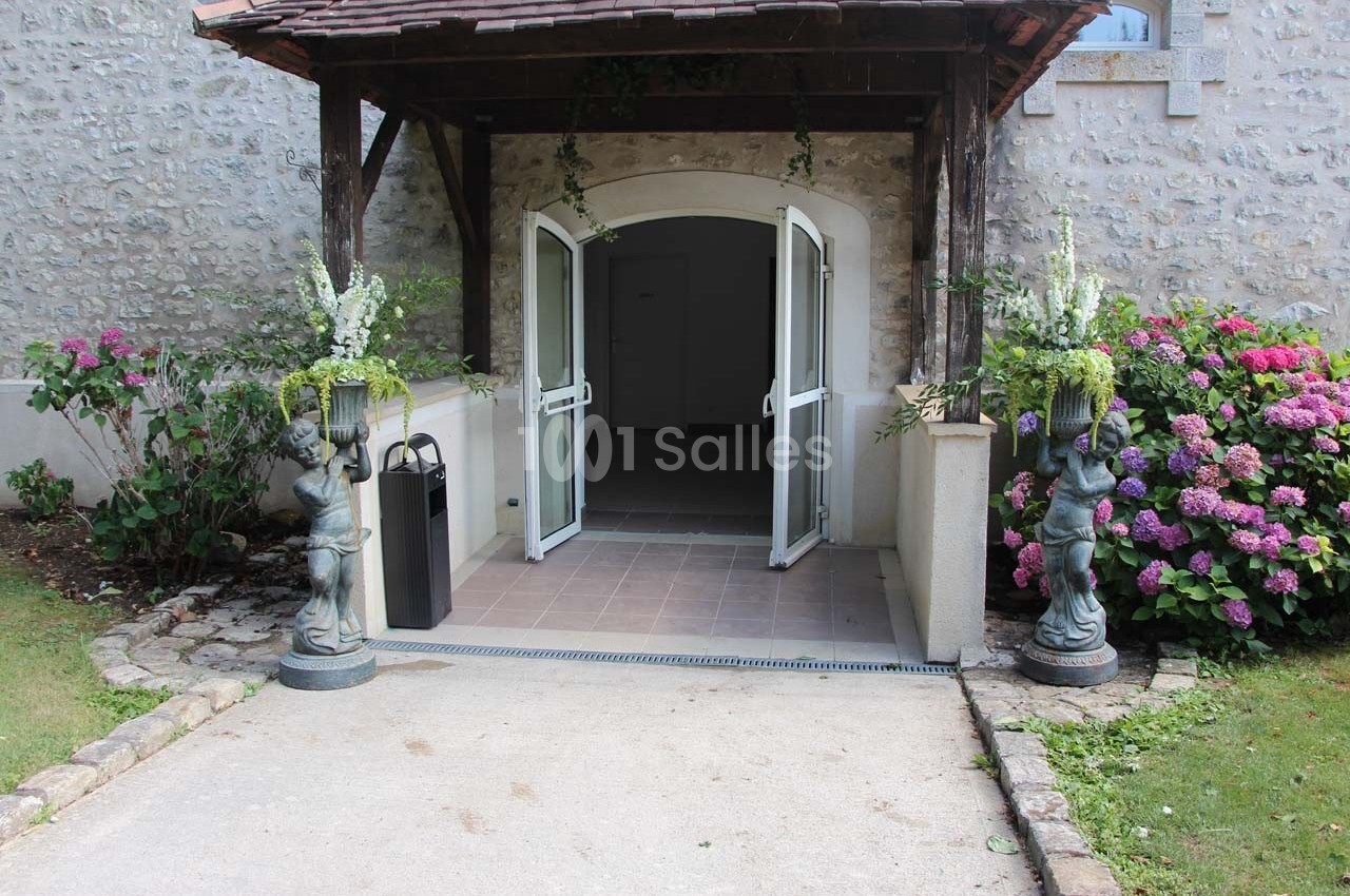 Entrée d'un bâtiment en pierre avec porte ouverte, encadrée de statues et de fleurs colorées.