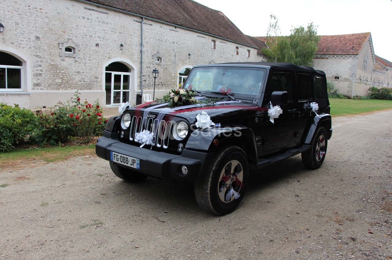 Voiture noire décorée de fleurs blanches et rouges pour un mariage, stationnée devant un bâtiment en pierre.