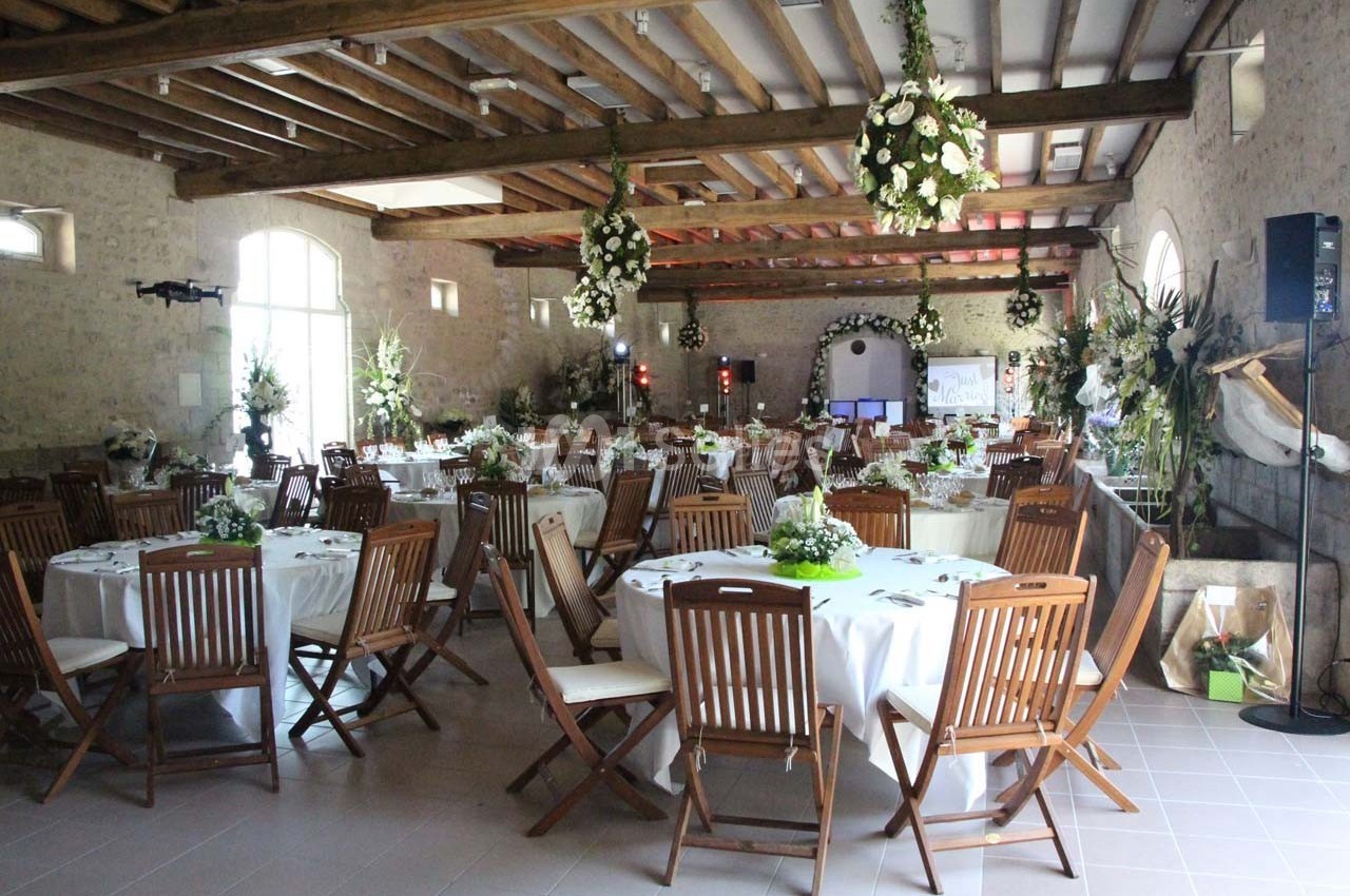 Salle de réception décorée avec des tables rondes, nappes blanches, chaises en bois et compositions florales suspendues.