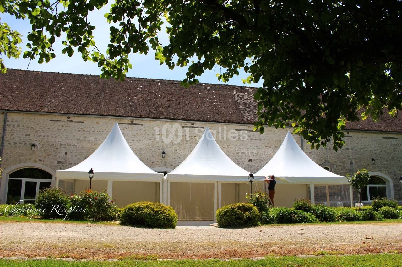 Trois tentes blanches installées devant un bâtiment en pierre, entourées de verdure et d'arbustes.