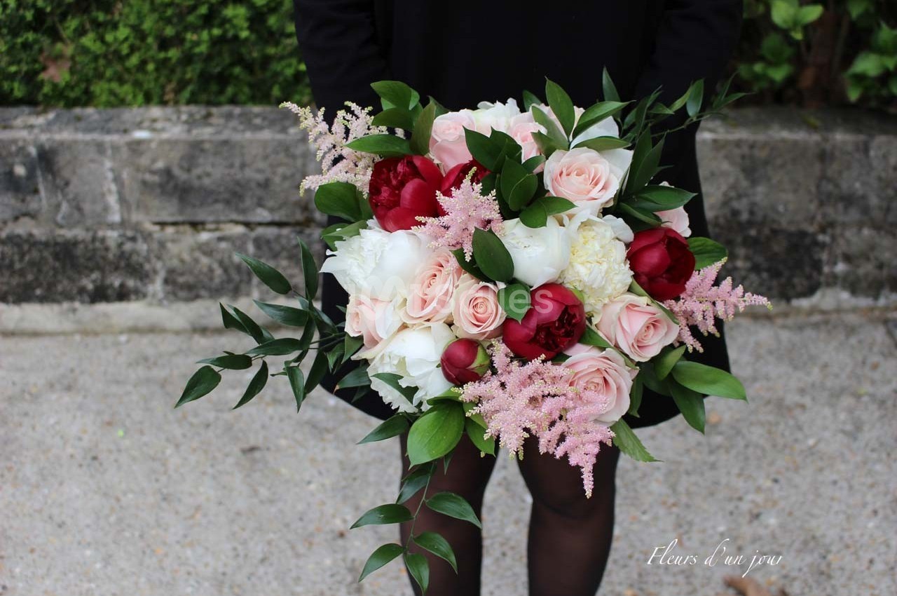 Bouquet de fleurs composé de roses, pivoines, astilbes roses et feuillage, tenu par une personne en arrière-plan flou.