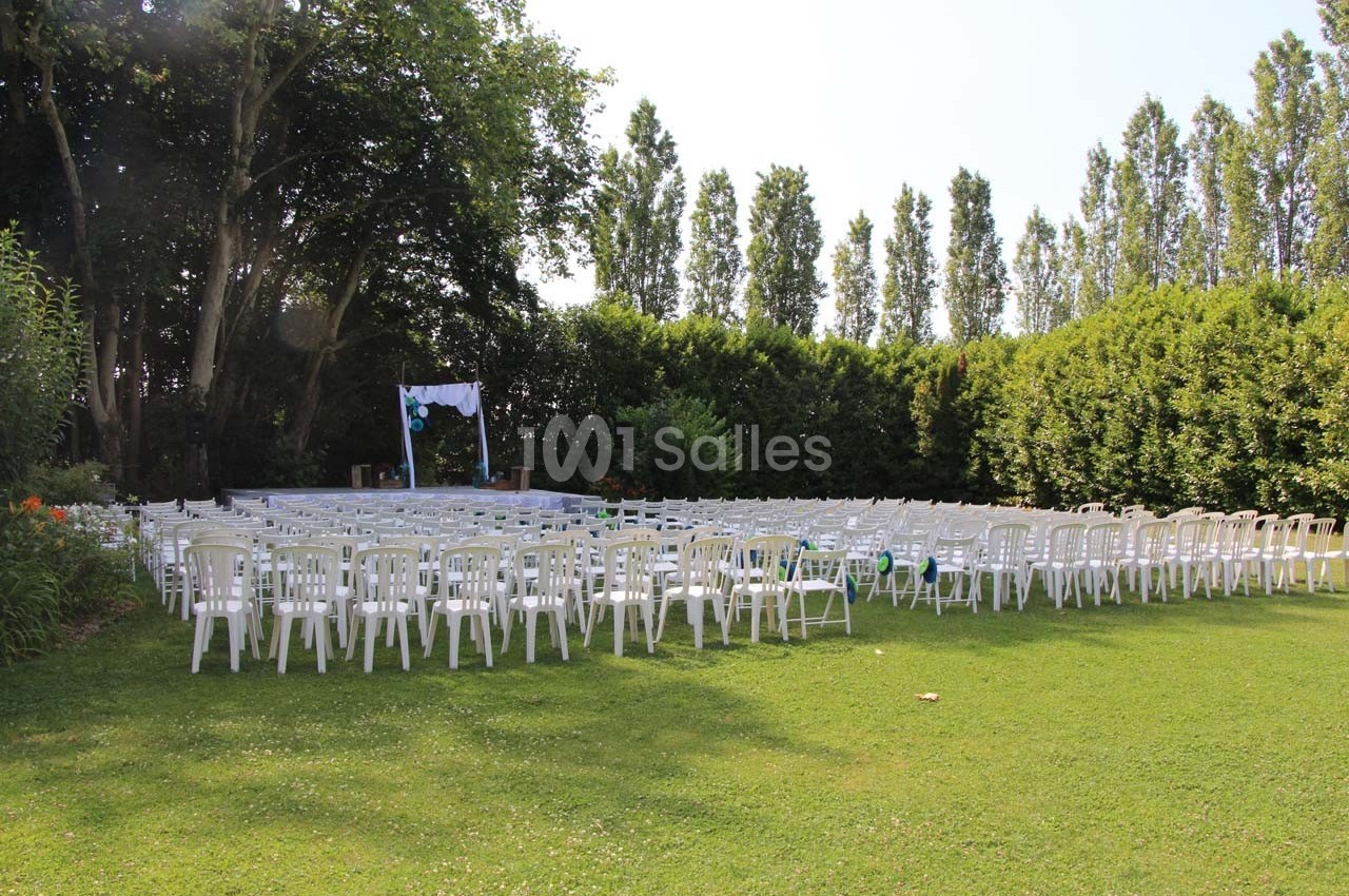 Chaises blanches disposées en rangées sur une pelouse, face à une petite scène décorée pour un événement en plein air.