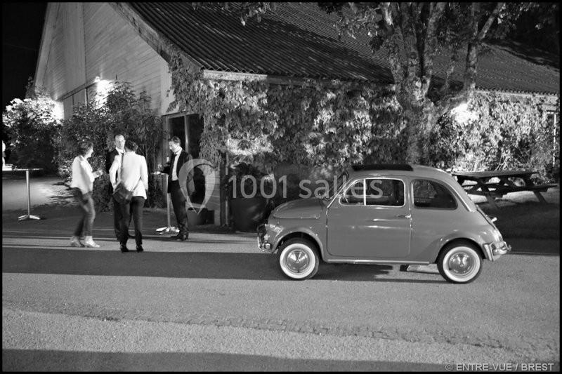 Une petite voiture ancienne garée devant un bâtiment éclairé, avec trois personnes discutant à proximité.