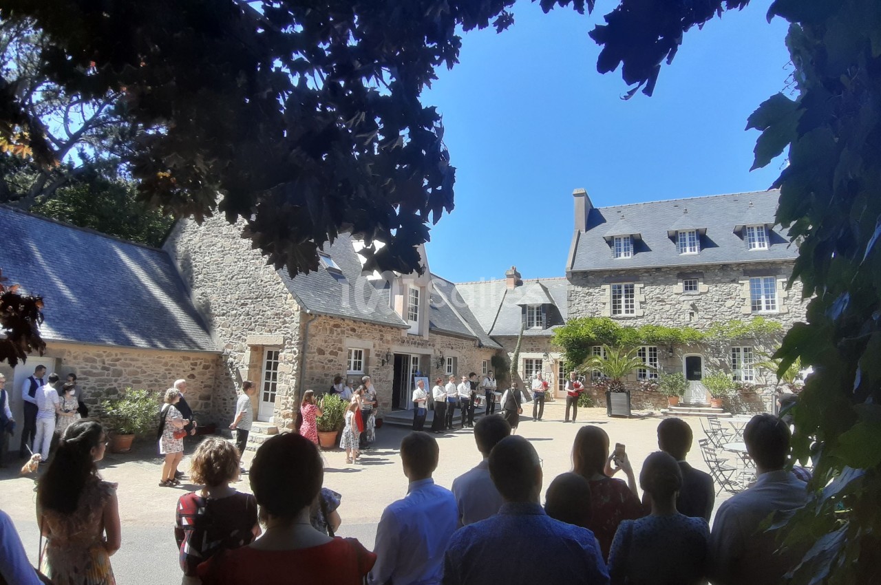 Groupe de personnes rassemblées dans une cour pavée entourée de bâtiments en pierre sous un ciel bleu clair.
