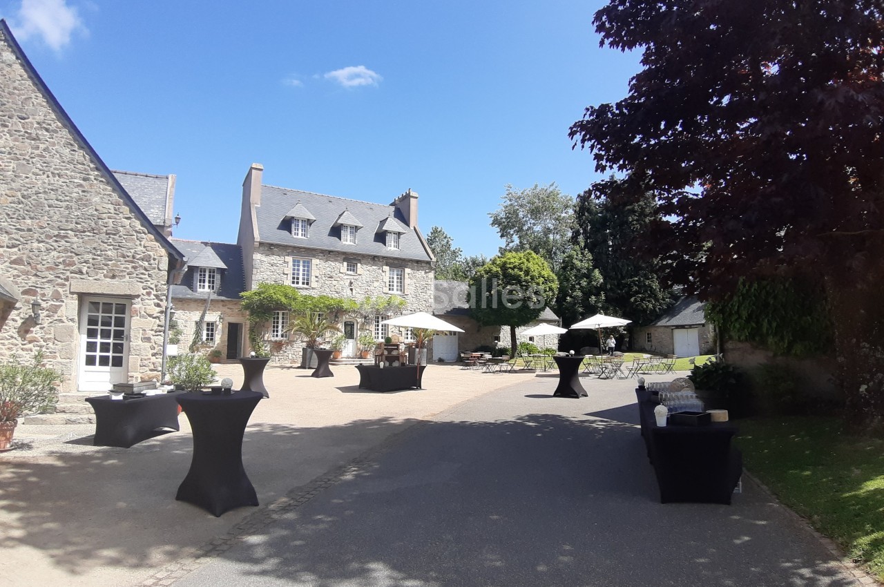 Cour d’un bâtiment en pierre avec tables hautes noires, parasols blancs et végétation sous un ciel ensoleillé.