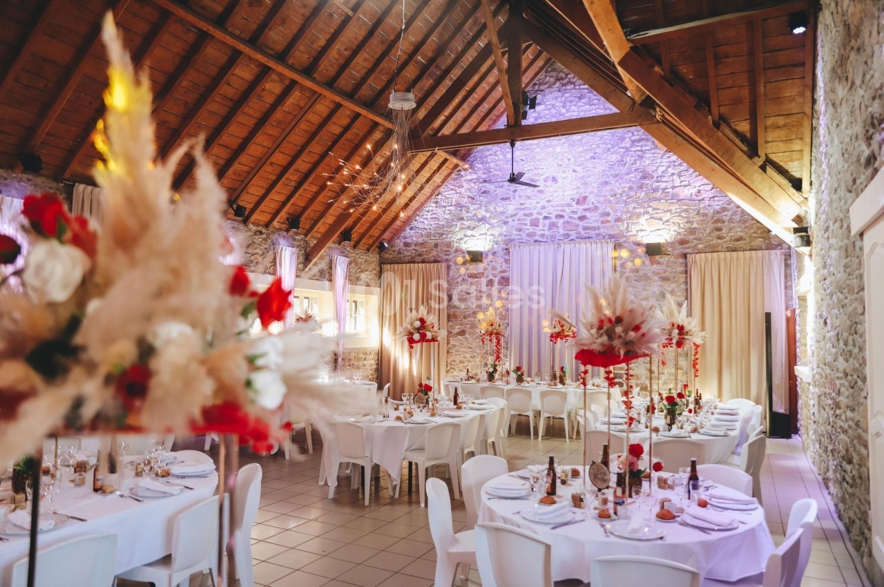 Salle de réception décorée avec des tables rondes, des chaises blanches et des compositions florales rouges et beiges.