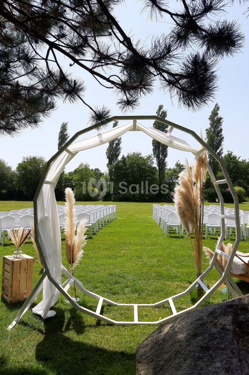 Arche circulaire décorée de pampas sur une pelouse, entourée de chaises blanches disposées pour une cérémonie en plein air.