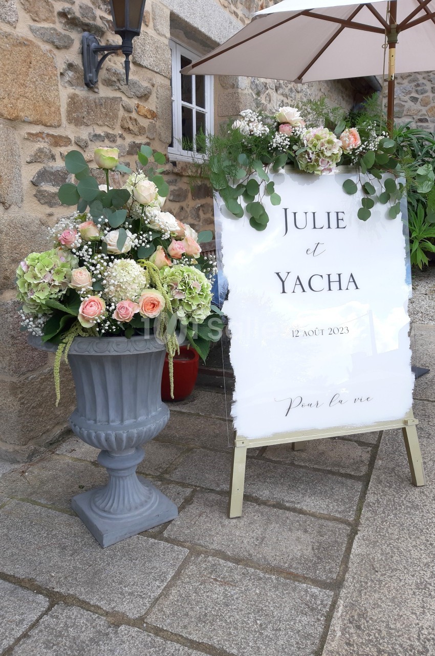Panneau de mariage décoré de fleurs blanches et roses, placé à côté d'une jardinière fleurie devant un bâtiment en pierre.