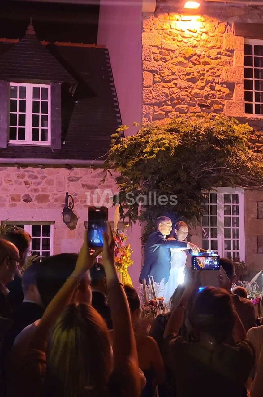 Un couple pose sous un éclairage nocturne devant une maison en pierre, entouré de personnes prenant des photos.
