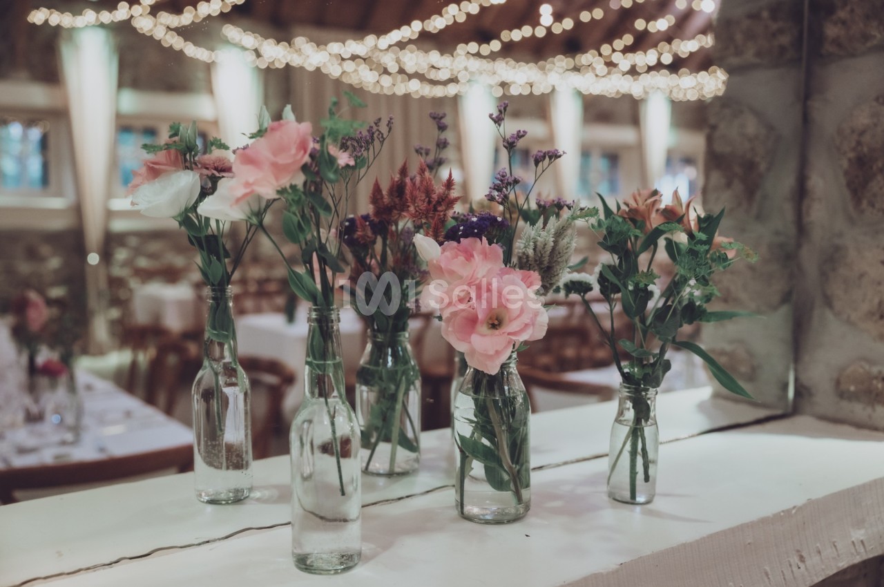 Bouquets de fleurs variées disposés dans des bouteilles en verre sur une table, avec des lumières suspendues en arrière-plan.