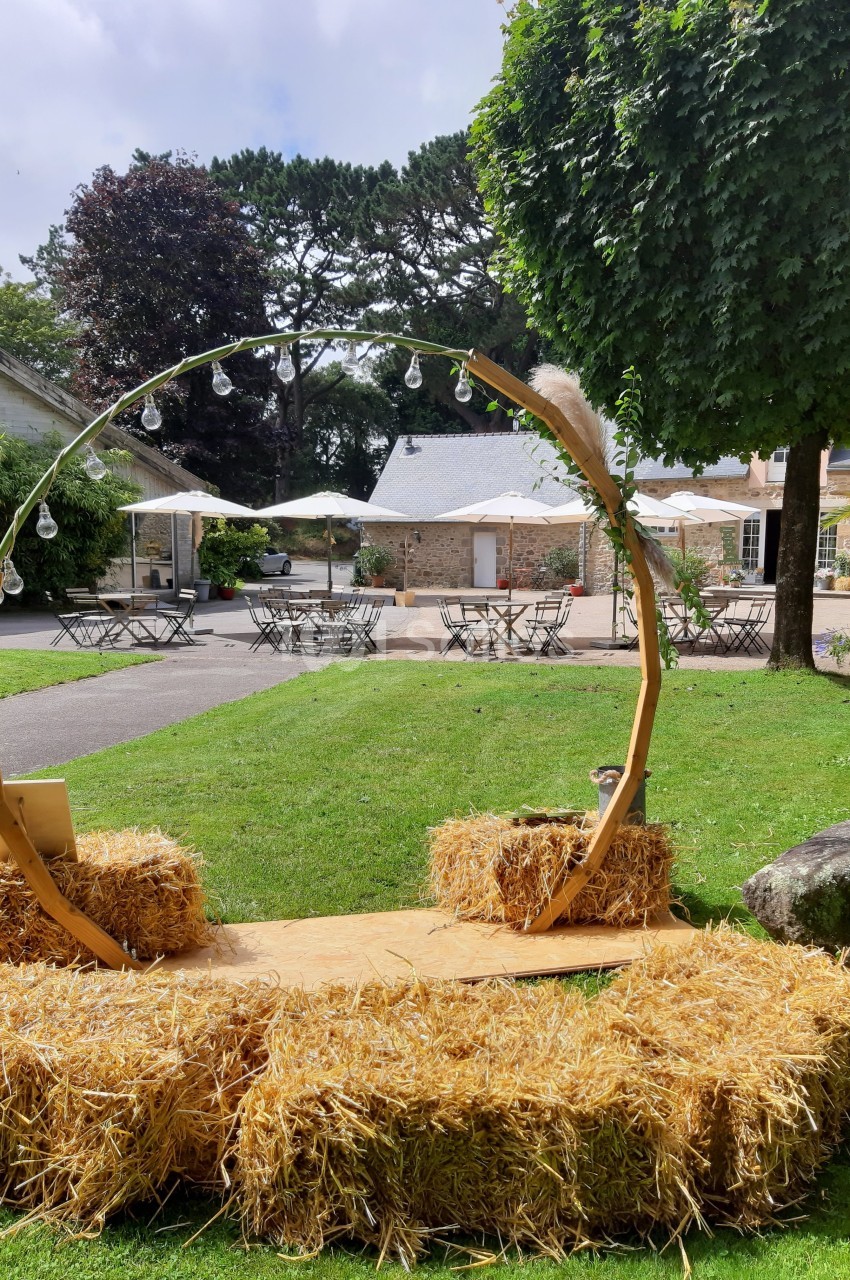 Arc décoratif en bois entouré de bottes de paille sur une pelouse, avec des tables et parasols en arrière-plan.