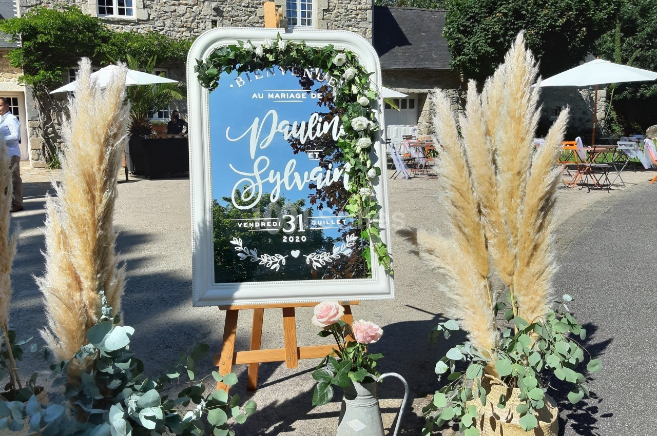 Panneau de mariage décoré de fleurs et de feuillages, placé devant une bâtisse en pierre avec des plantes ornementales.