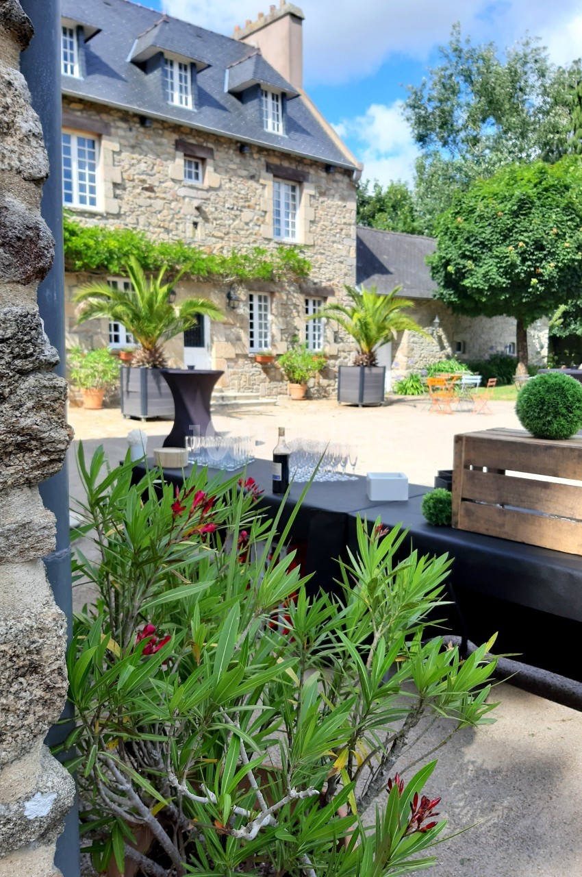 Cour d'une maison en pierre avec des plantes, des tables hautes et des caisses en bois, sous un ciel partiellement nuageux.