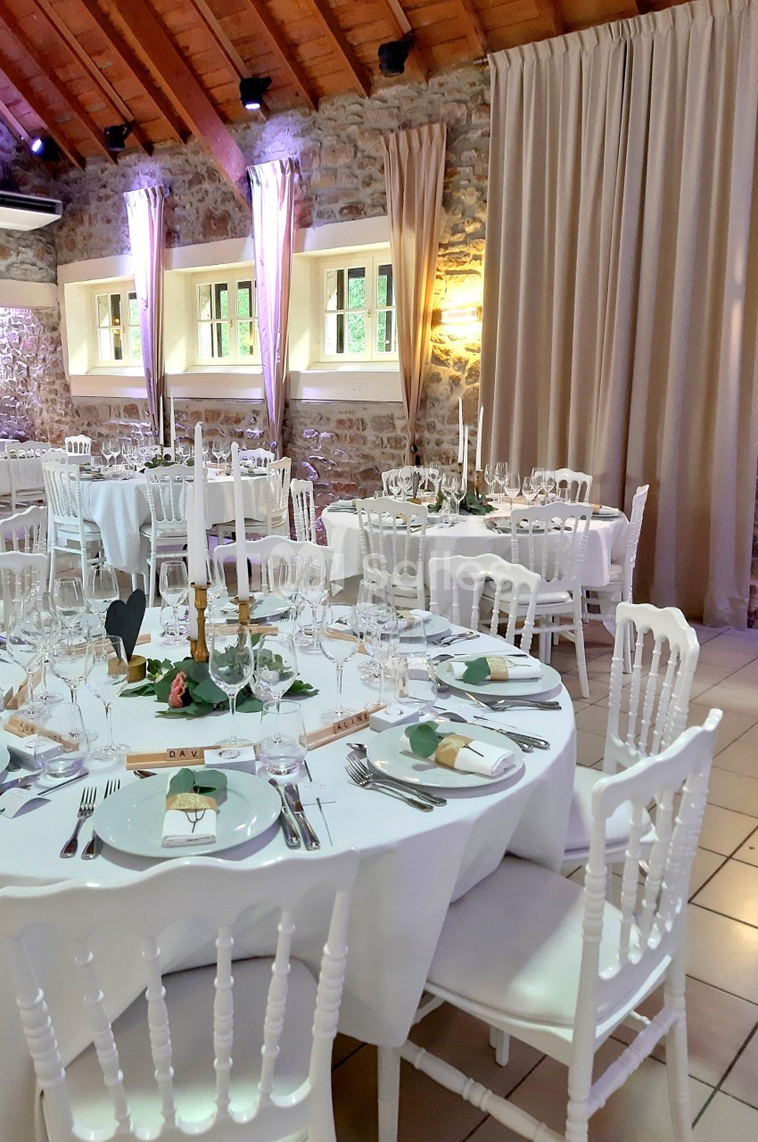 Salle de réception décorée avec des tables rondes dressées, nappes blanches, chandeliers et murs en pierre apparente.