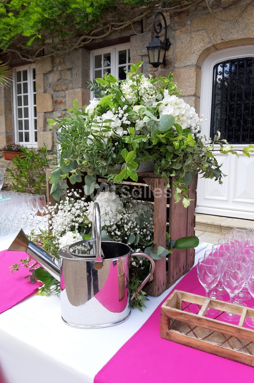 Arrangement floral avec des fleurs blanches et feuillages dans une caisse en bois, arrosoir en métal et verres en arrière…