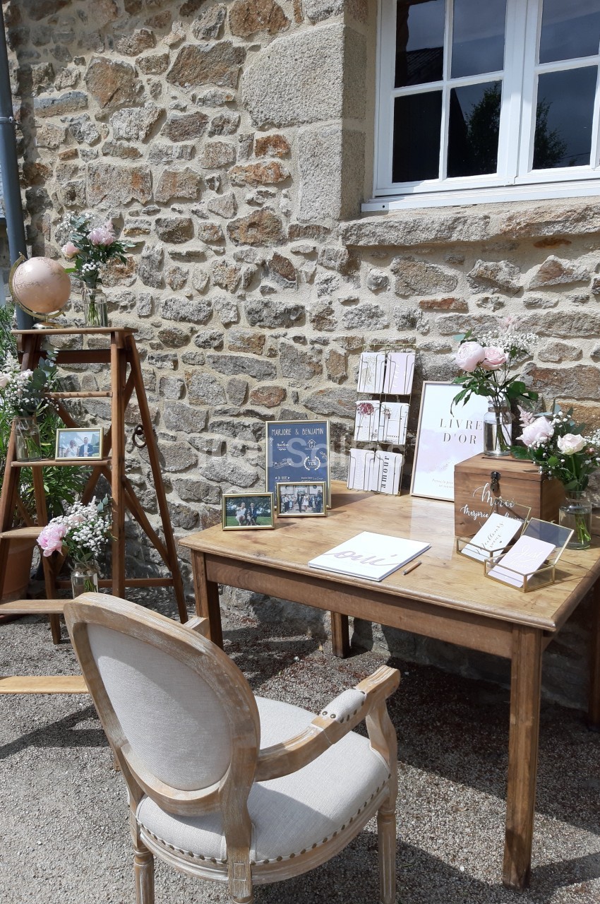 Table en bois avec livre d'or, décorations florales et échelle ornée de cadres, devant un mur en pierre.