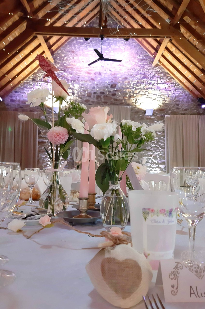 Table de réception décorée avec des fleurs, bougies roses et vaisselle élégante dans une salle en pierre avec poutres…