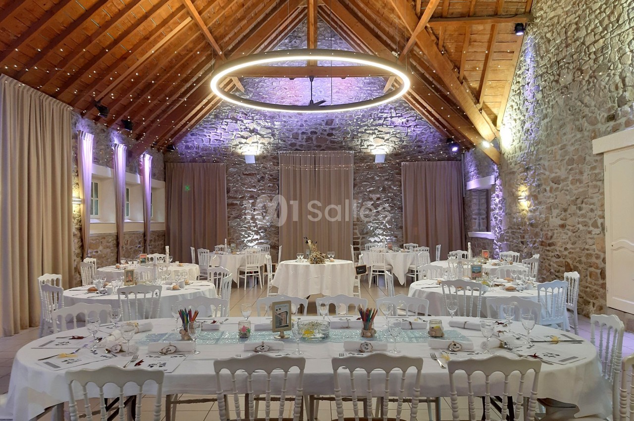 Salle de réception en pierre avec tables décorées, chaises blanches et éclairage chaleureux sous un plafond en bois.
