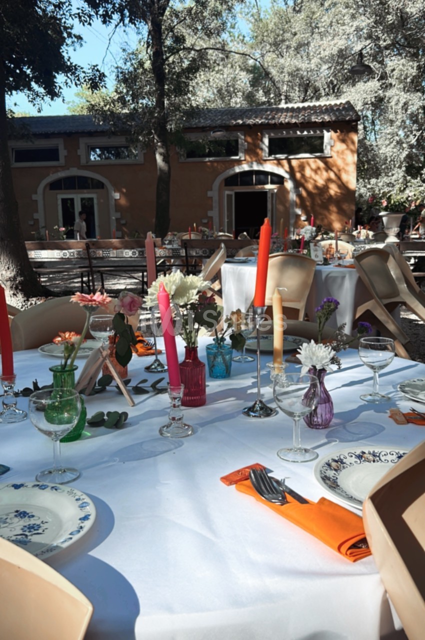 Table dressée en extérieur avec nappes blanches, bougies colorées, vaisselle et décorations florales sous des arbres.