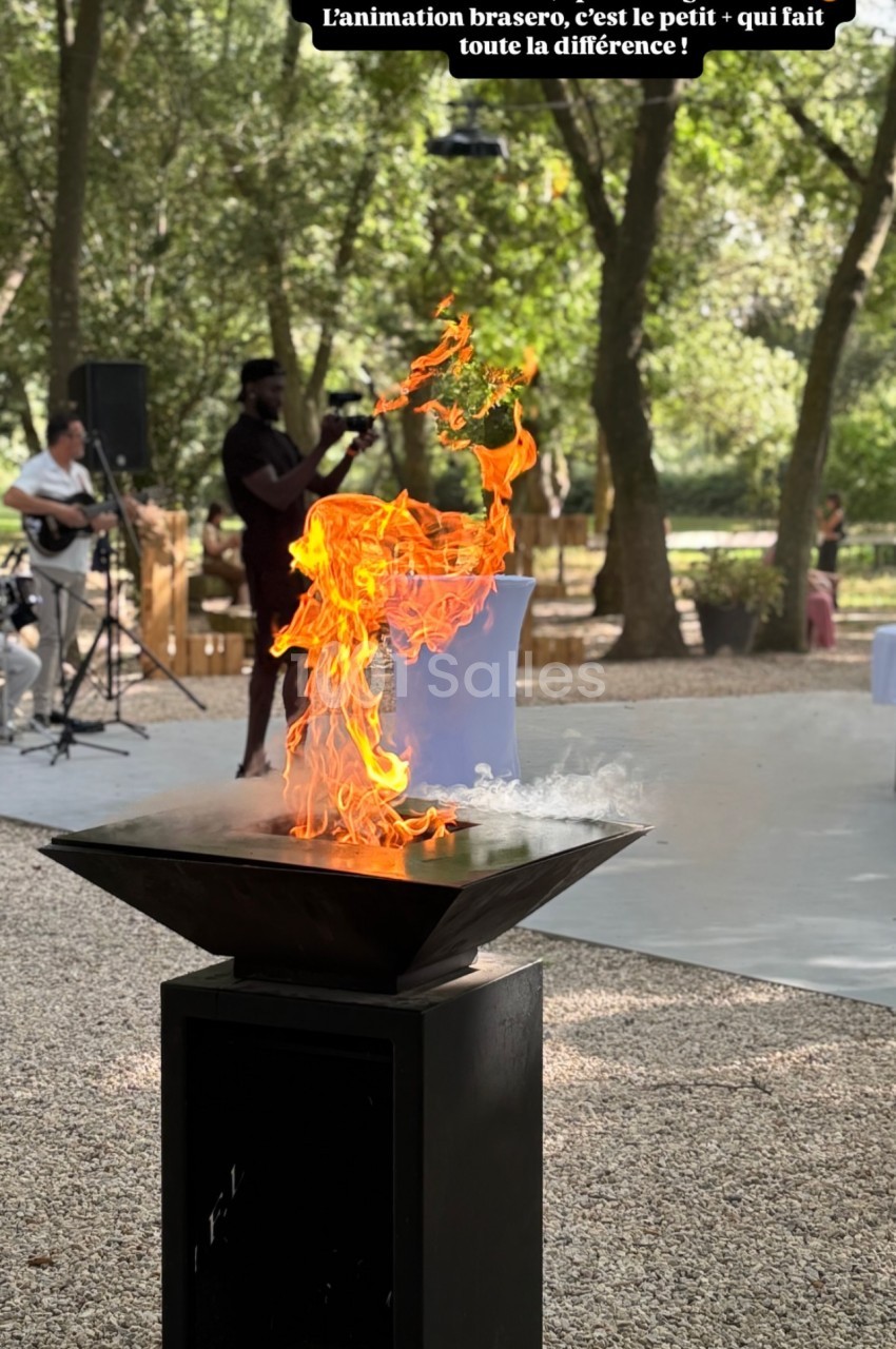Brasero allumé avec des flammes vives dans un parc arboré, un musicien en arrière-plan.