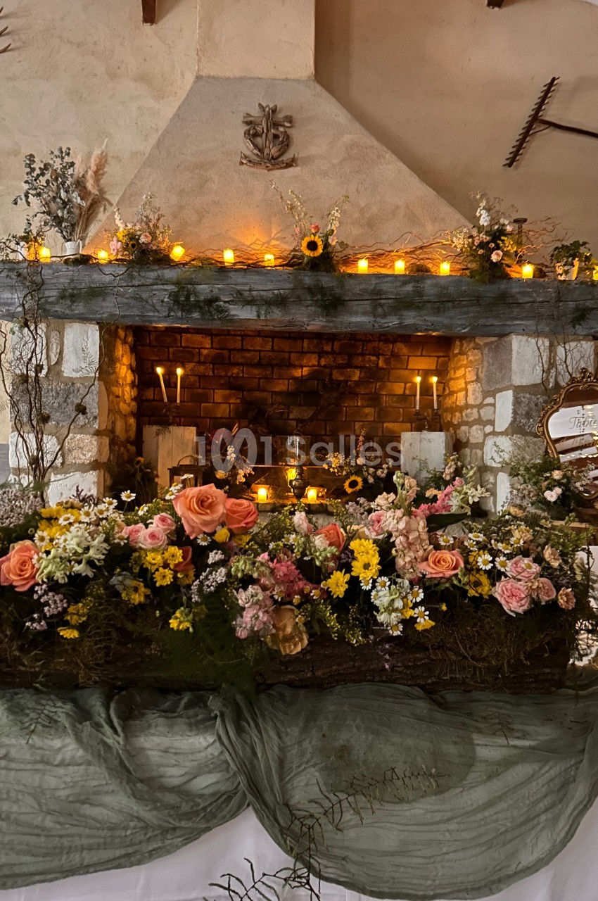 Cheminée décorée de fleurs colorées, bougies allumées et guirlande lumineuse dans un cadre rustique.