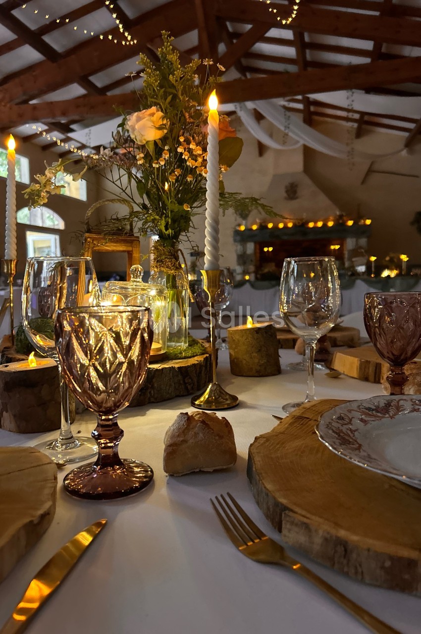 Table décorée pour un dîner, avec bougies allumées, verres colorés, rondins de bois et fleurs dans une salle chaleureuse.