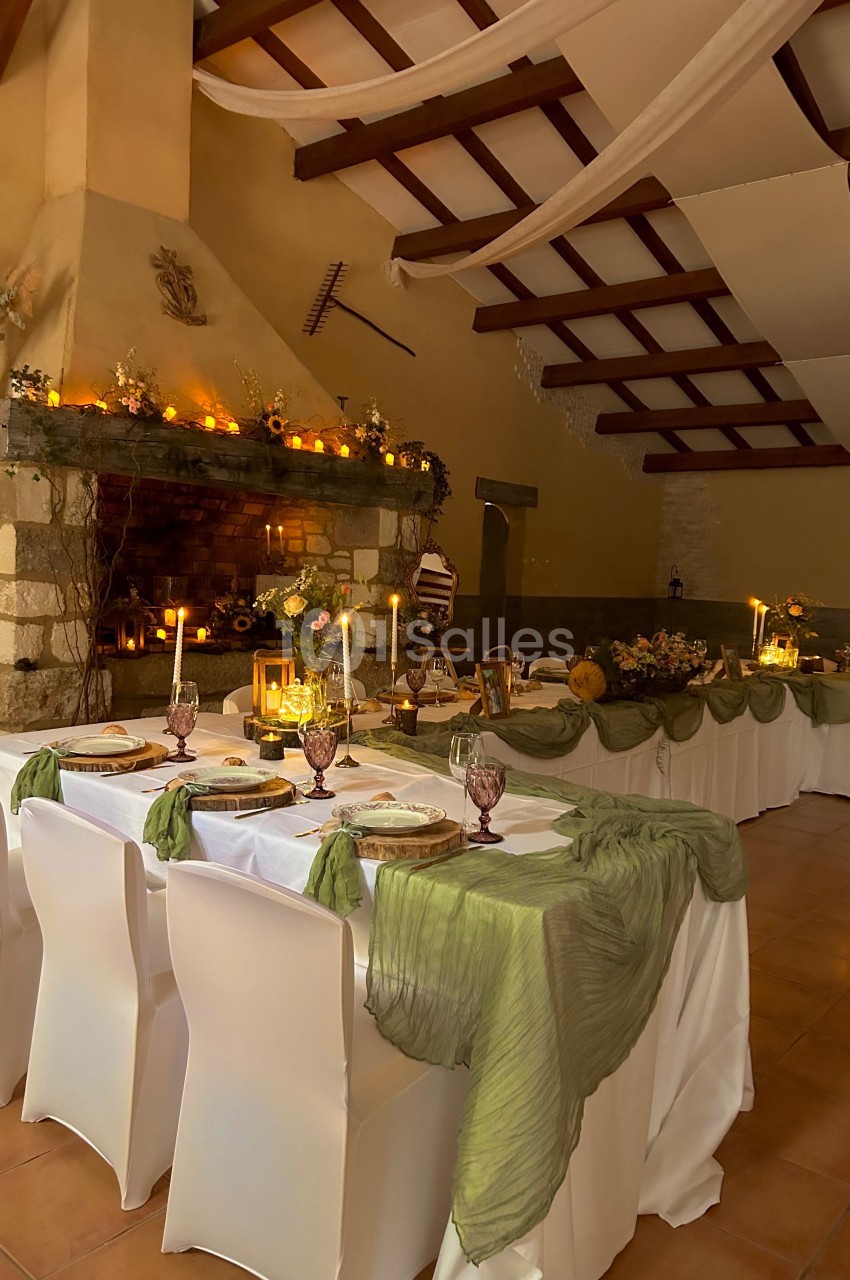 Salle décorée pour un repas, avec tables dressées, nappes blanches, voilages verts et cheminée ornée de bougies.