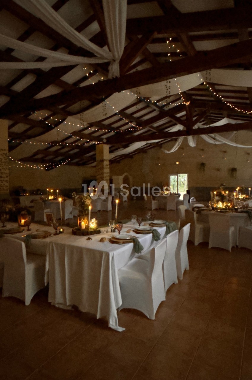Salle de réception décorée avec des guirlandes lumineuses, des tables dressées et des chaises recouvertes de housses…