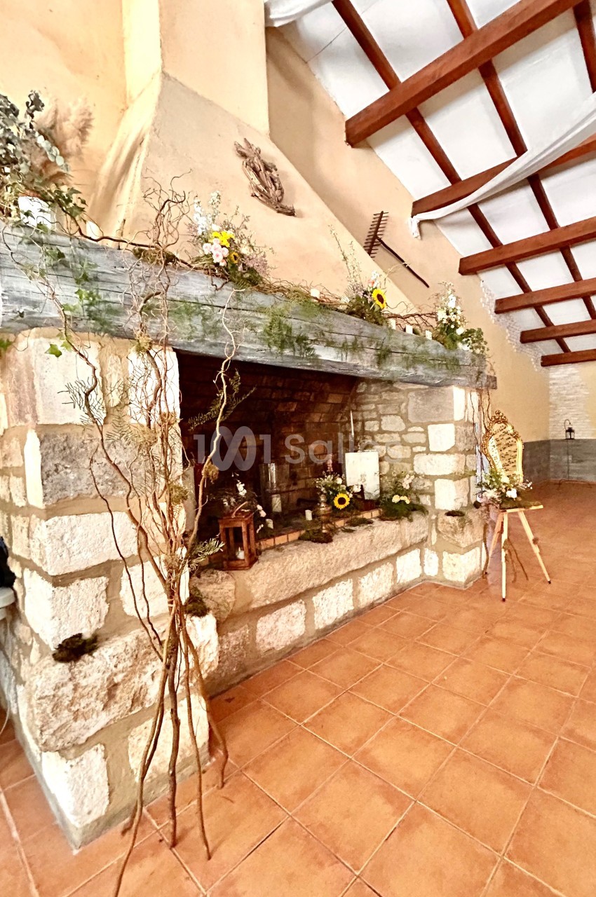 Cheminée en pierre décorée de fleurs et branchages dans une pièce avec plafond en poutres apparentes.