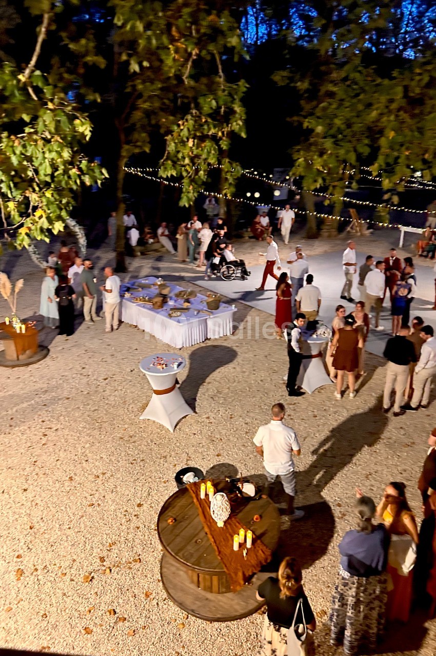 Groupe de personnes réunies en soirée en plein air, avec tables décorées et guirlandes lumineuses suspendues.