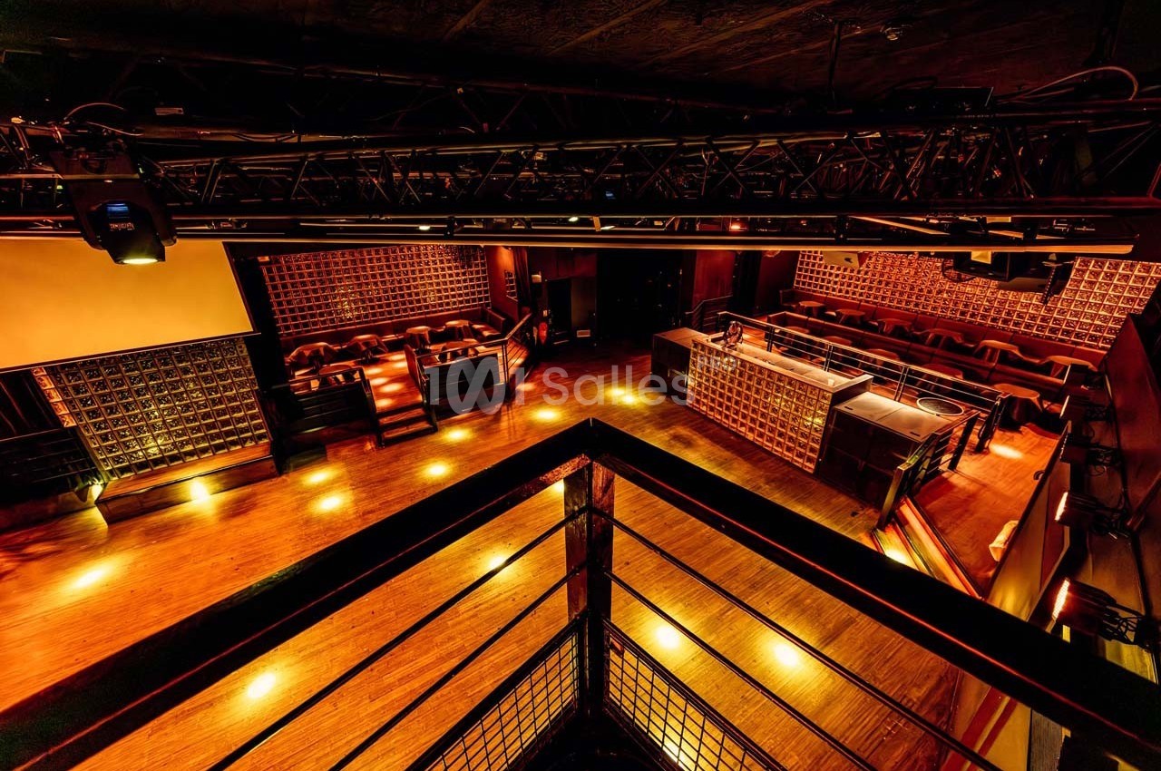 Salle de spectacle vide avec éclairage chaleureux, vue depuis une mezzanine sur la scène et les sièges en contrebas.