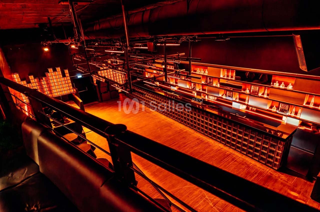 Salle intérieure sombre avec un bar éclairé par des lumières orangées, vue depuis une mezzanine avec garde-corps.