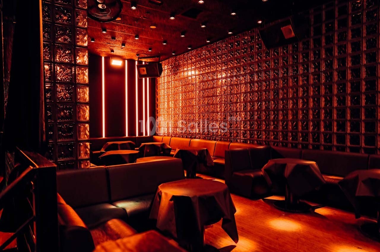 Salle sombre avec murs en briques de verre, éclairage rouge tamisé et tables recouvertes de nappes noires.