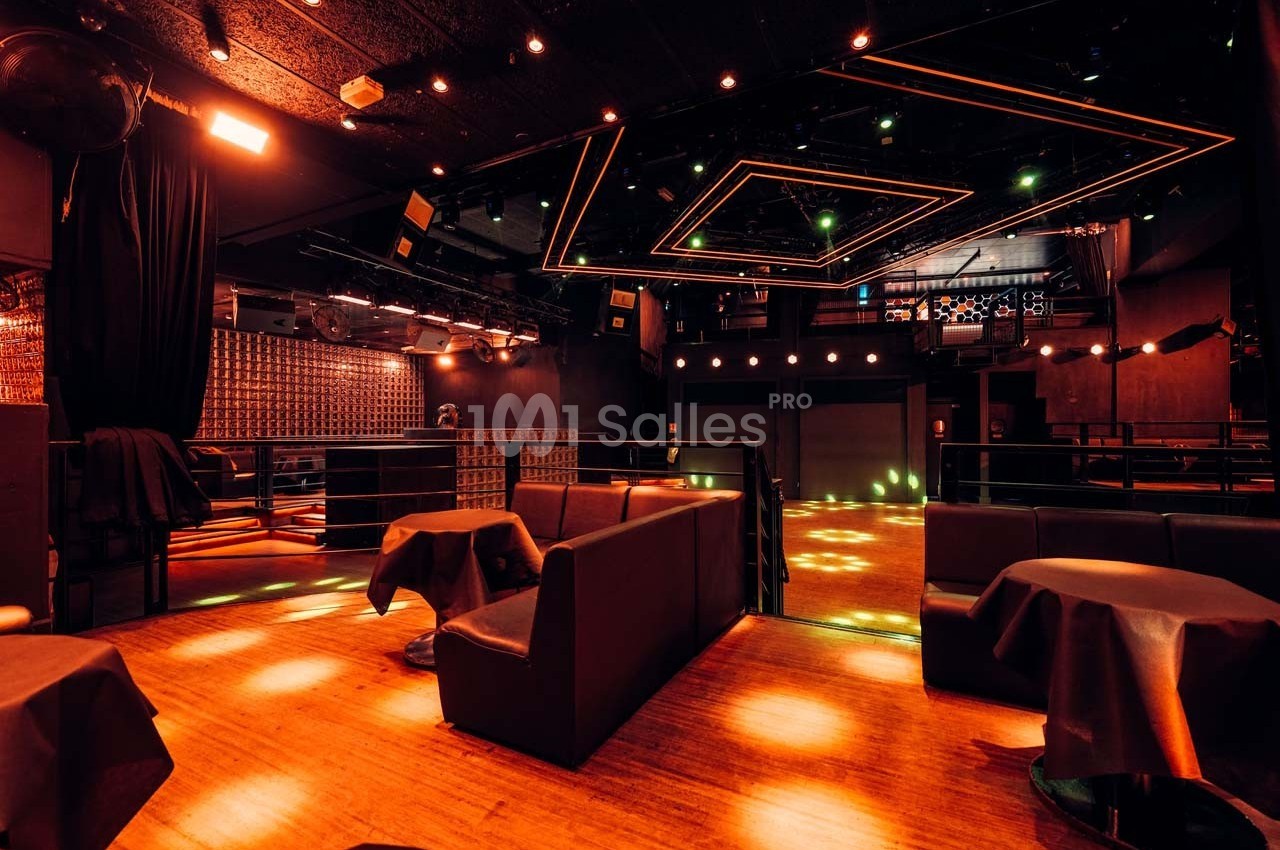 Salle de spectacle sombre avec éclairage tamisé, tables et banquettes noires, et piste de danse illuminée.