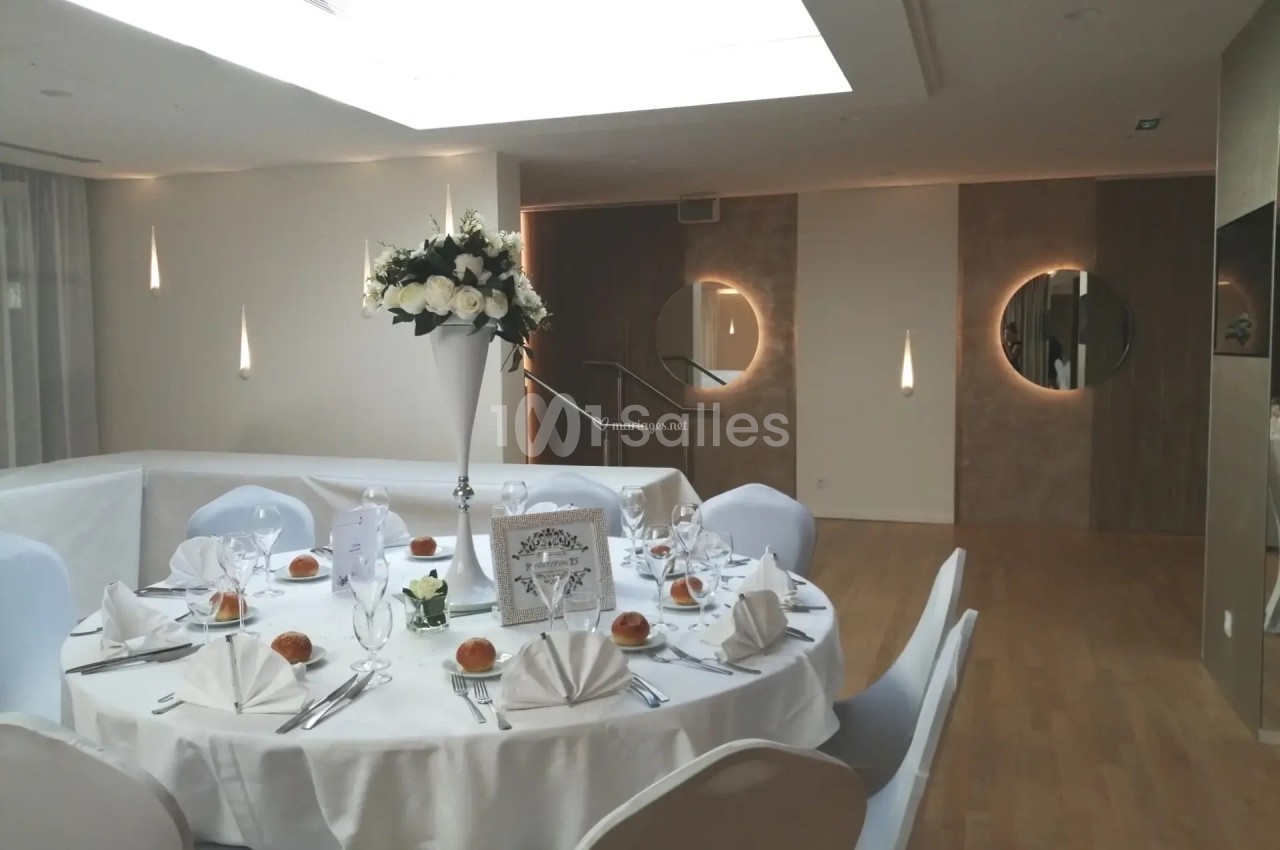 Salle de réception élégante avec table ronde dressée, nappes blanches, serviettes pliées et centre de table floral.