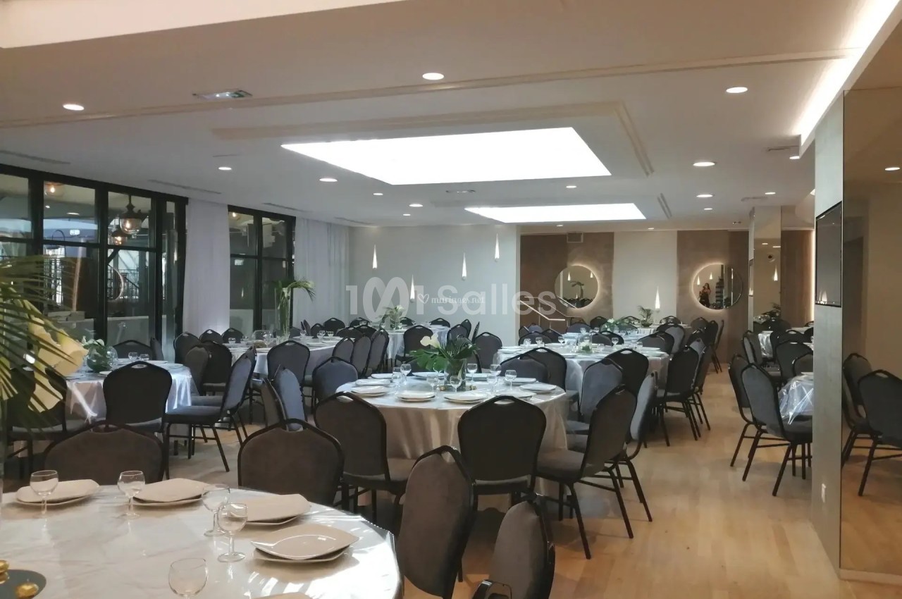 Salle de réception lumineuse avec tables rondes dressées, chaises noires et décoration sobre.