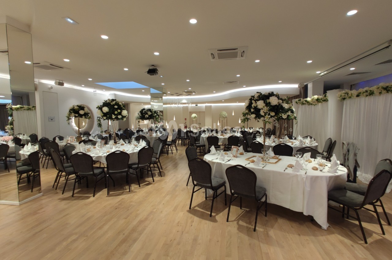 Salle de réception élégante avec tables rondes dressées, décorations florales blanches et éclairage chaleureux.