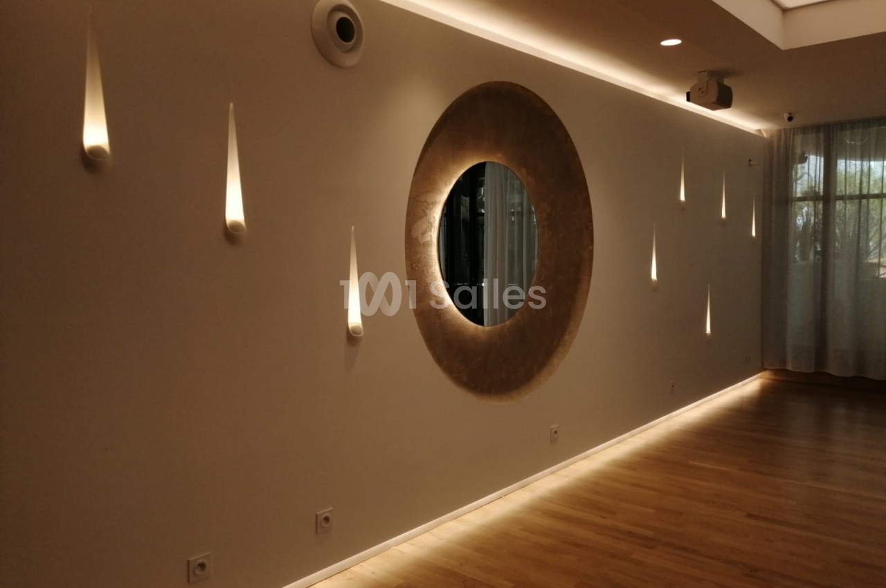 Mur beige avec un miroir rond central, éclairages muraux en forme de gouttes et sol en parquet clair.