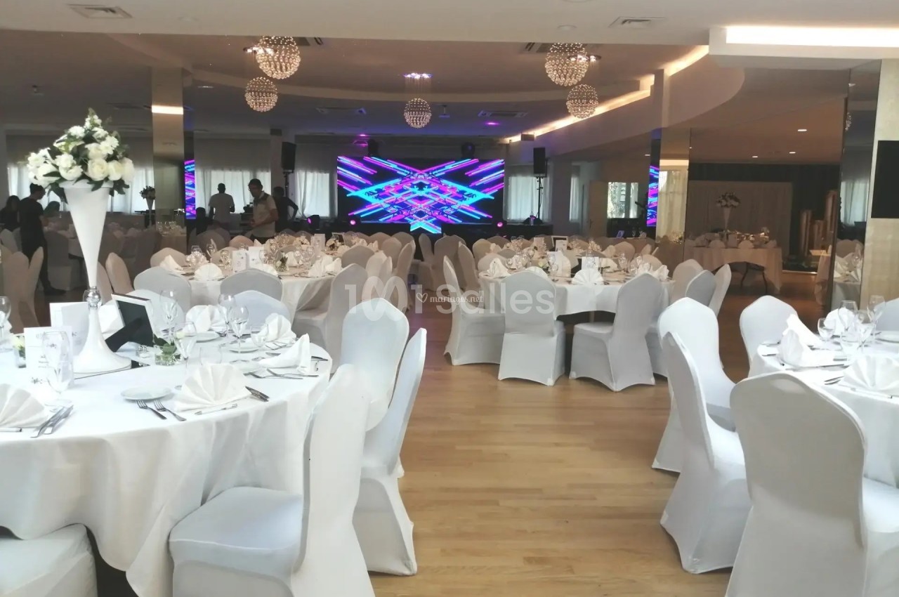 Salle de réception élégante avec tables rondes dressées, chaises blanches et écran lumineux en arrière-plan.