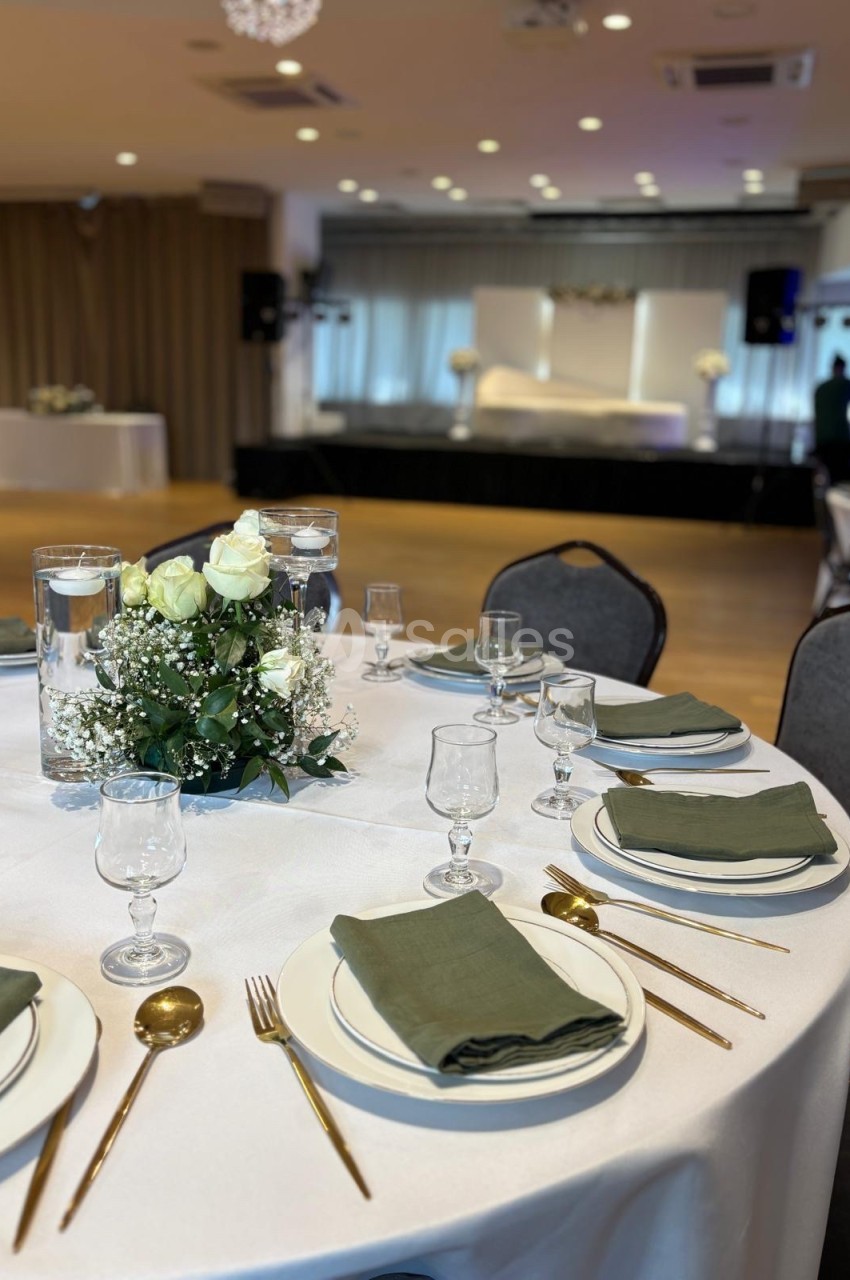 Table dressée avec une nappe blanche, couverts dorés, serviettes vertes et un centre de table floral dans une salle de…