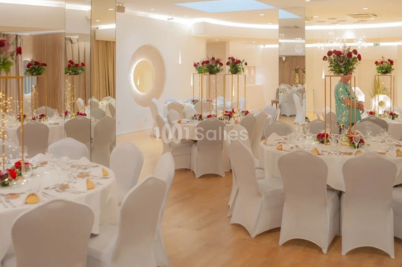 Salle de réception élégante avec tables rondes dressées, nappes blanches et décorations florales rouges et blanches.