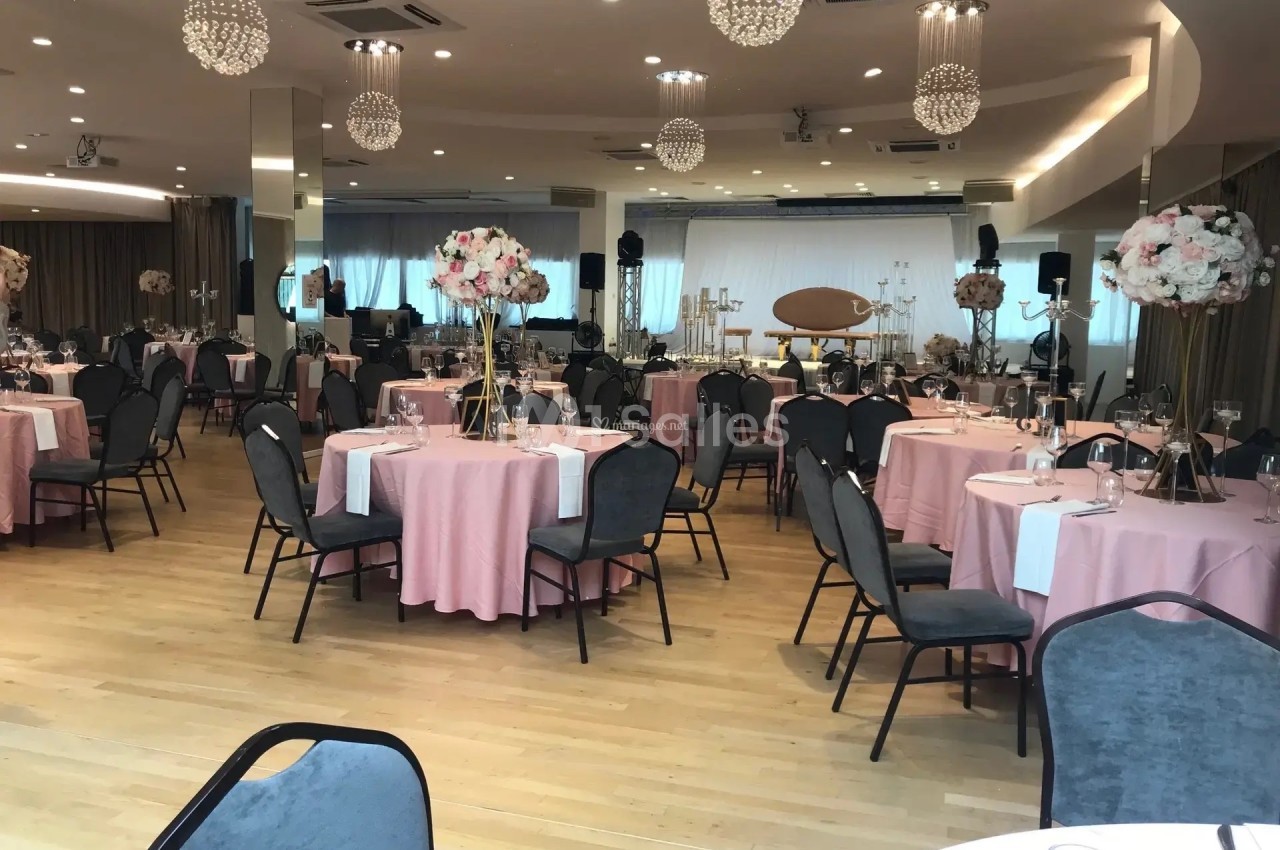 Salle de réception élégante avec tables rondes dressées, nappes roses et décorations florales sur supports dorés.