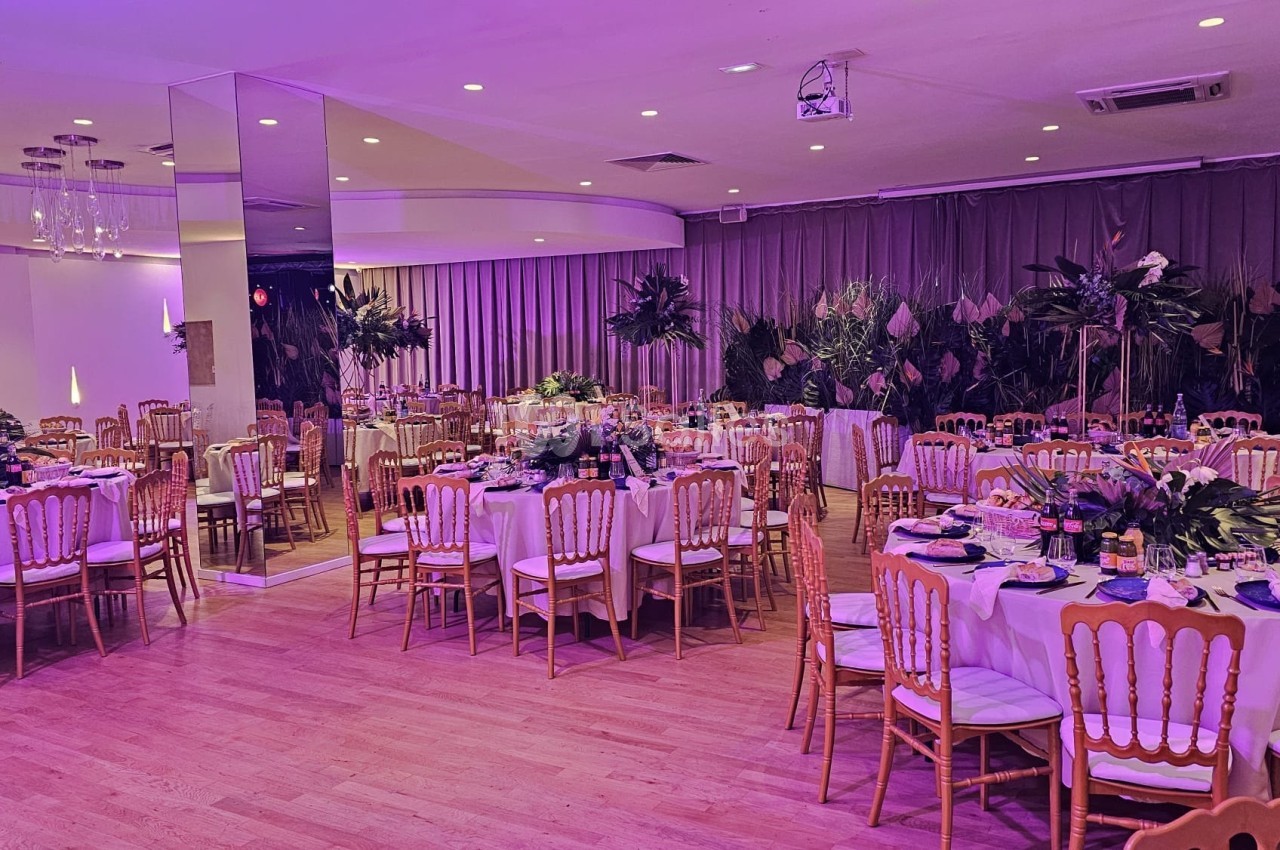 Salle de réception décorée avec des tables rondes dressées, des chaises en bois et un éclairage violet tamisé.