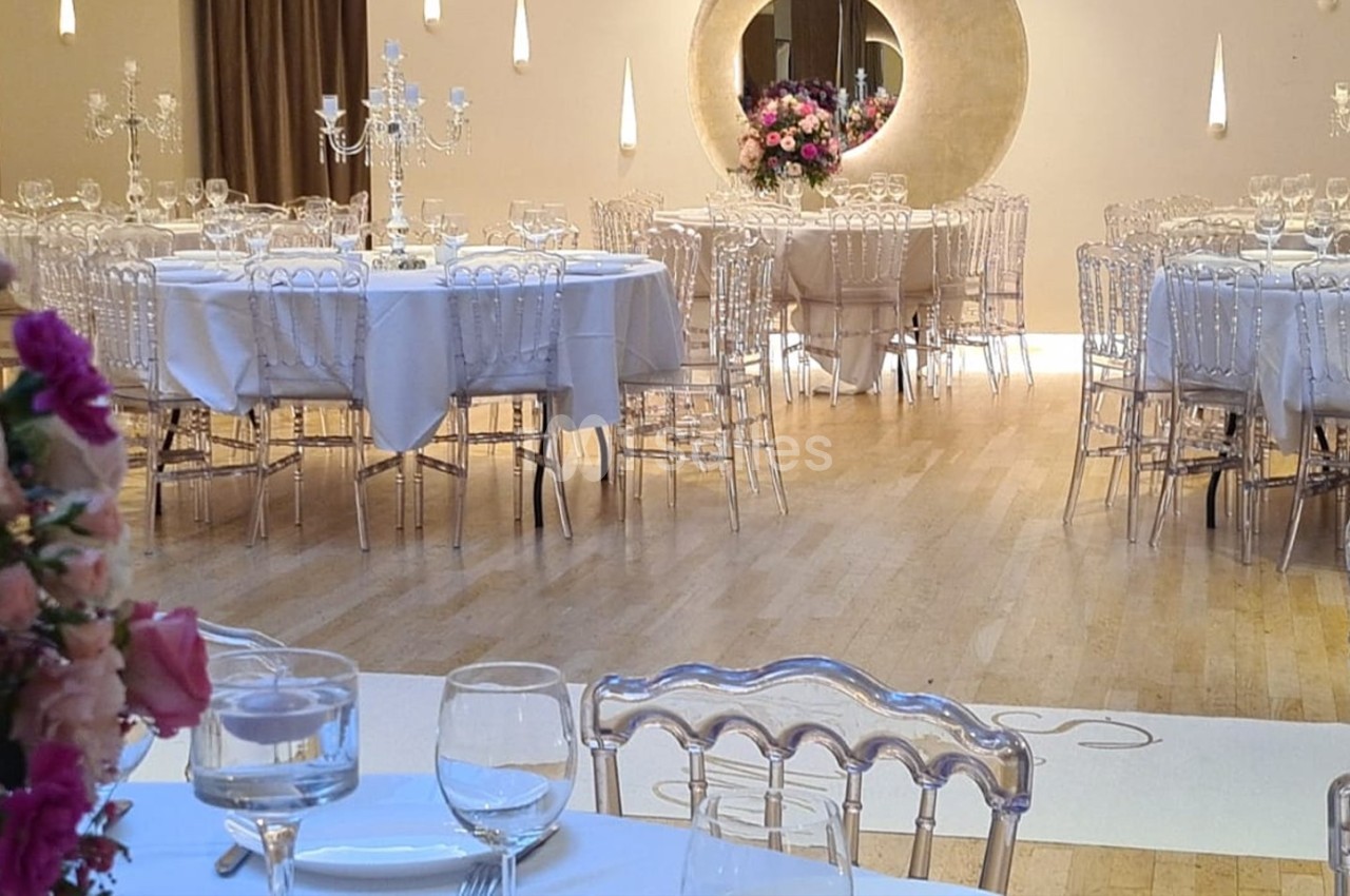 Salle de réception élégante avec tables rondes dressées, chaises transparentes et décorations florales.